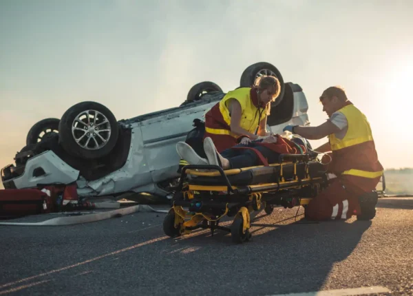 On the site of a car crash, two paramedics tend to a victim on a stretcher in front of a turned over vehicle.