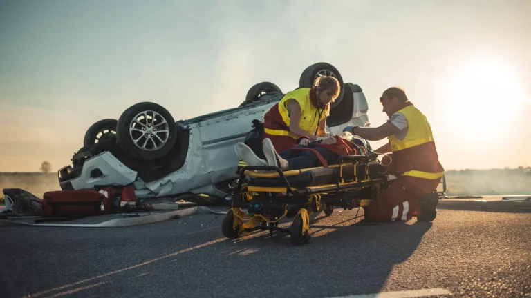 On the site of a car crash, two paramedics tend to a victim on a stretcher in front of a turned over vehicle.