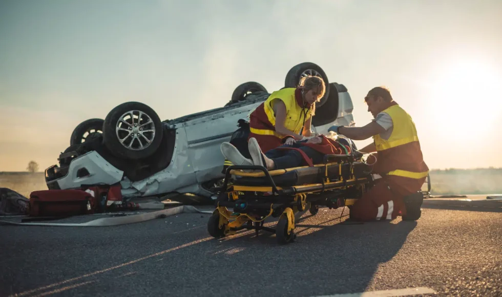 On the site of a car crash, two paramedics tend to a victim on a stretcher in front of a turned over vehicle.