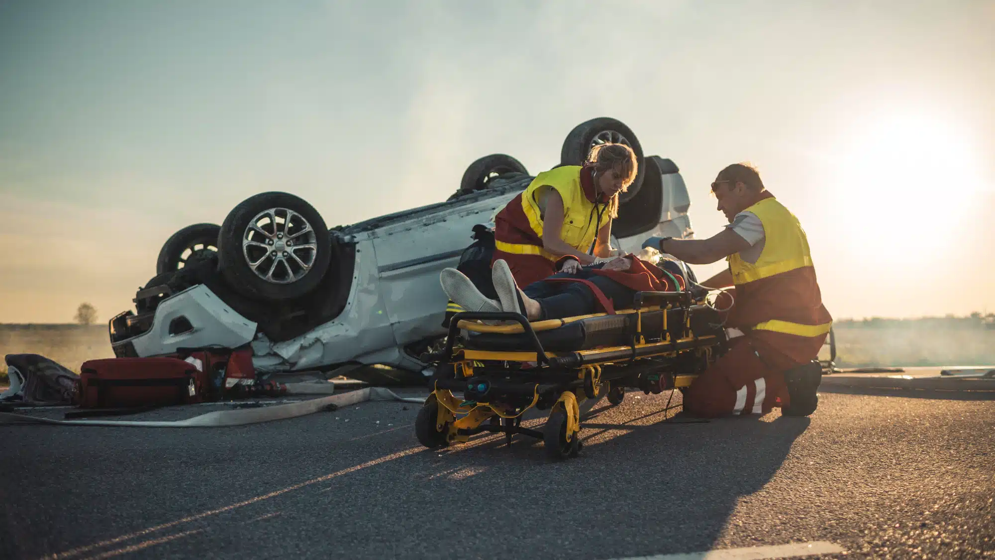 On the site of a car crash, two paramedics tend to a victim on a stretcher in front of a turned over vehicle.